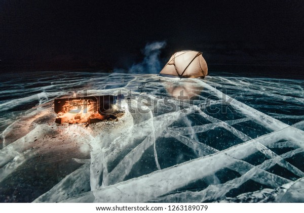 Fire on ice at night. Campground on ice. Tent stands\
next to bonfire. Lake Baikal. Nearby there is car. Shelter tent and\
ice are illuminated from the inside. Beautiful bonfire on real\
cracked ice