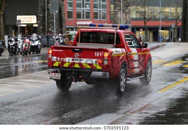 Fire service car photographed during a rainy spring day\
Lausanne Switzerland, March 2020. Red fire car is driving in the\
urban area of city. In the background you can see some scooters and\
buildings. 