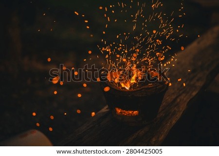 Fire spark flame ember charcoal brazier heat campfire night outdoor glow from burning charcoal in metal bowl on wooden surface with flying orange spark and dark rustic mood