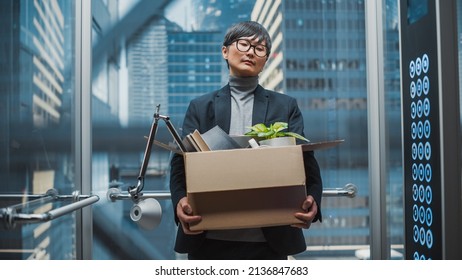 Fired Manager Going Down From Office In A Glass Elevator In Modern Business Center. Sad Specialist Laid Off, Feeling Depressed And Devastated, Holding Office Accessories And A Plant In A Box.