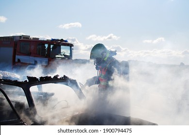 A Firefighter With A Breathing Apparatus Extinguishes A Car Fire Against The Rays Of The Sun With Smoke And Water Splinters In The Background