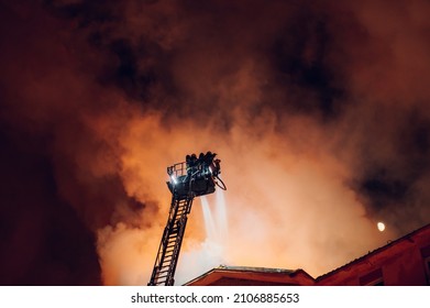 Firefighter directed hose towards the burning building at night. Boom crane with firefighters. Firefighter on pull-out crane. Firefighter extinguishes fire.