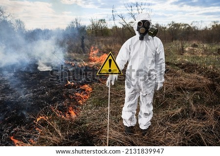 Firefighter ecologist extinguishing fire in field. Man in protective radiation suit and gas mask near burning grass with smoke, holding warning sign with exclamation mark. Natural disaster concept.