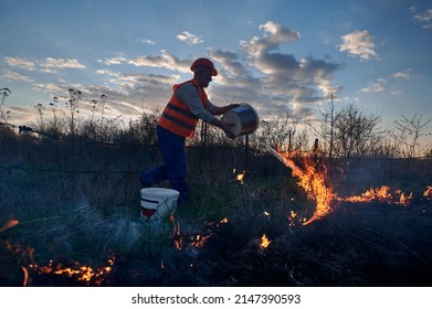 Firefighter Ecologist Fighting Wildfire In Field With Evening Sky On Background. Male Environmentalist Holding Bucket And Pouring Water On Burning Dry Grass. Natural Disaster Concept.