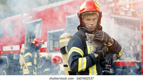 Firefighter in fire fighting operation. Portrait of heroic fireman in protective suit and red helmet holds saved cat in his arms, second fireman is out of focus near fire engine