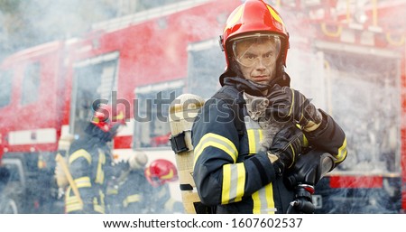 Firefighter in fire fighting operation. Portrait of heroic fireman in protective suit and red helmet holds saved cat in his arms, second fireman is out of focus near fire engine