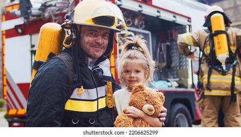 Firefighter Hug Rescued Little Girl With Teddy Bear. Frightened Child Rejoices In Rescue. At Background Firefighters After Extinguishing Fire Next To Fire Truck. Concept Of Saving Lives, Fire Safety