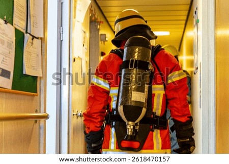 A firefighter, seen from behind,  in full protective gear, mask, and oxygen tank stands ready during a fire drill on board. Fully equipped, he executes fire suppression tasks with required precision.