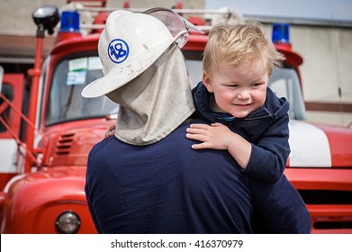 A Firefighter Take A Little Child Boy To Save Him. Fire Engine Car On Background. Fireman With Kid In His Arms. Protection Concept