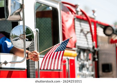 A firefighter waves an American flag from the driver's seat window of a firetruck during a patriotic US parade for Memorial Day, Flag Day, or July 4th.