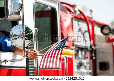 A firefighter waves an American flag from the driver's seat window of a firetruck during a patriotic US parade for Memorial Day, Flag Day, or July 4th.