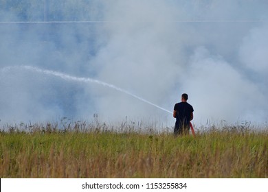 Firefighter Without Helmet And Protective Equipment Holding Red Hose With Water And Spraying Stream On Smoke Of Burning Wild Fire In Green Meadow In Hot Summer Day.
