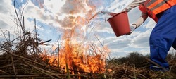 Fireman Ecologist Extinguishing Fire In Field With Cloudy Sky On Background. Close Up Of Man Holding Bucket And Pouring Water On Burning Dry Grass. Natural Disaster And Ecology Demage Concept.
