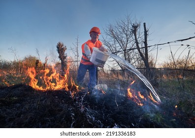 Fireman Ecologist Extinguishing Fire In Field With Evening Sky On Background. Male Environmentalist Holding Bucket And Pouring Water On Burning Dry Grass. Natural Disaster Concept.