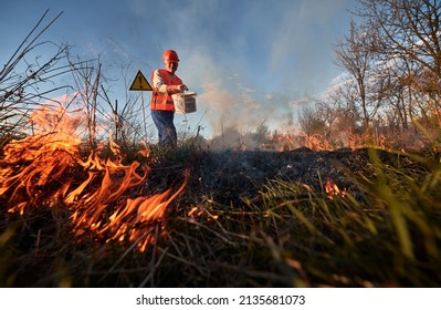 Fireman ecologist fighting fire in field with evening sky on background. Low angle view man holding bucket and pouring water on burning dry grass near warning sign with exclamation mark.