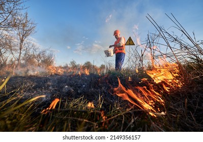 Fireman ecologist fighting wildfire in field with evening sky on background. Male environmentalist holding bucket and pouring water on burning dry grass near warning sign with exclamation mark.