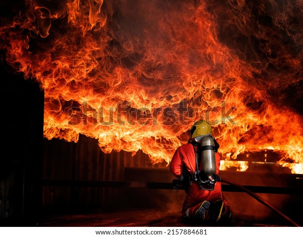 Fireman or firefighter from fire wearing fire\
protection suit and helmet and breathing with oxygen tank hold the\
water hoses spraying water to extinguish the heavy fire burn and\
smoke overhead