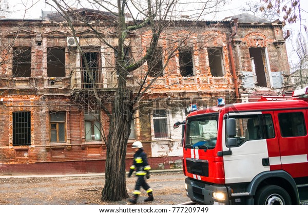 The fireman walks next to the fire engine against\
the ruined building. A fire truck is standing next to the emergency\
house. Selective focus