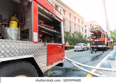 Firemen Wearing Uniform Standing Behind Fire Tape And Many Fire Engine Trucks With Ladder At Accident In Highrise Tower Residential Or Office Building In City Center. Emergency Rescue At Disaster