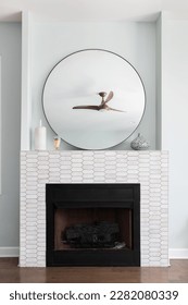 A fireplace detail with marble picket tiles around the hearth, decorations and a large circular mirror above, and hardwood flooring.