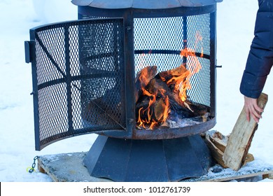 Fireplace In Winter Season Charged With Firewood, In The Burning Process. Wood-burning Fire Pit With Removable Fire Pan And With Wide Bar Ribs And Handles. Outdoor Chimney With Steel Grille And Cap