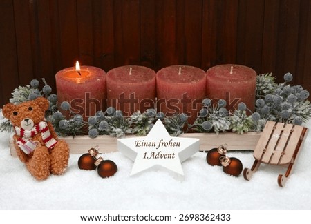 First Advent: Rust-colored Advent candles decorated with a teddy bear, Christmas baubles, and a sled in the snow. A star bears the inscription 