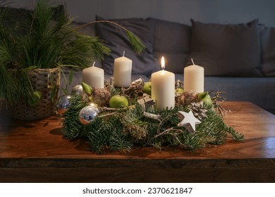 First Advent, wreath with one burning white candle and Christmas decoration on a wooden table in front of the couch, festive home decor, copy space, selected focus, narrow depth of field