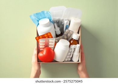 First aid kit containing essential medical supplies like pills, bandages, a thermometer, a face mask, and a red heart, held by two hands against a light green background