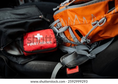 A first aid kit is placed right next to an orangecolored backpack, which is typically used for carrying personal belongings and essentials while traveling, hiking, or going about daily activities