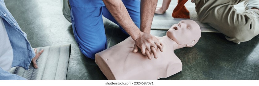 first aid seminar, hands-on learning, cropped view of paramedic showing chest compressions on CPR manikin near young participants in training room, life-saving skills and techniques concept, banner - Shutterstock ID 2324587091