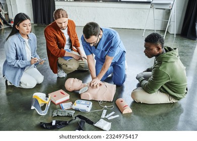 first aid seminar, multiethnic team looking at medical instructor doing chest compressions on CPR manikin near defibrillator, wound care simulators, neck brace, bandages and compression tourniquets