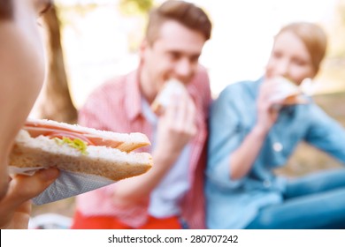 First Bite.  Close Up Of Young Girl Eating Sandwich On Background Of Another People During Picnic