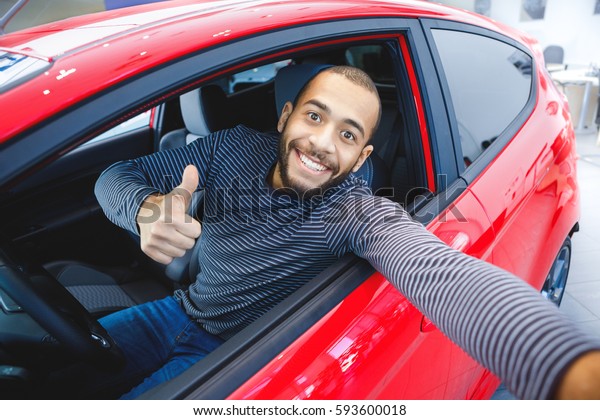 First car pic.\
Handsome young African man smiling making a selfie sitting in his\
new car at the dealership copyspace owner ownership driver. buyer\
consumerism lifestyle travel\
concept