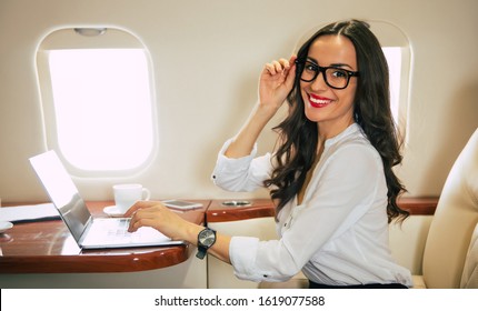 First class private jet. Close-up photo of a charming woman in a white blouse, who is typing something on her laptop, smiling and touching the frame of her glasses in an aircraft.