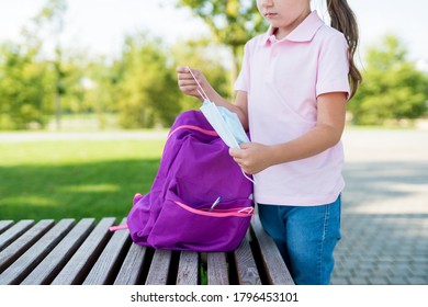 First Day Of School After Quarantine. Child Schoolgirl Student Staying Near The School And Taking Out  Protective Maskout Of The Her Backpack. Concept Back To School During Coronavirus Epidemic