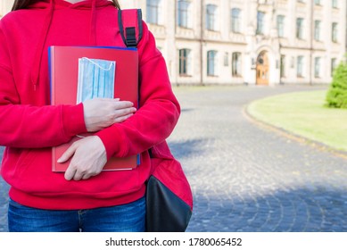 First Day At School After University Concept. Cropped Close Up Photo Of Teen Girl In Casual Red Sweater Holding Books And Medical Mask Standing Near Doors Enter