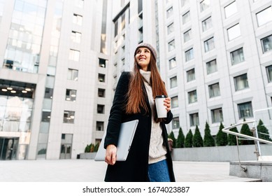 First Day At Work. A Young Woman With A Laptop And Coffee In Her Hands Is Standing Near The Office Building And Smiling.