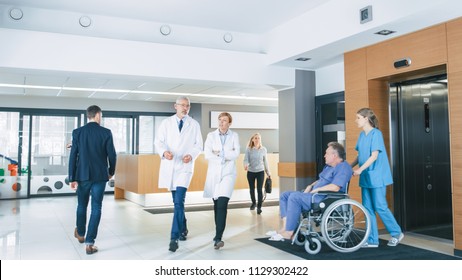 First Floor Of The Busy Hospital, Doctors, Nurses And Personnel Busy Working, Assistant Moves Elderly Man In The Wheelchair. New Modern Medical Hospital With Professional Staff.