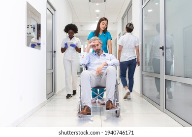 First Floor Of The Busy Hospital, Doctors, Nurses And Personnel Busy Working, Assistant Moves Elderly Man In The Wheelchair. New Modern Medical Hospital With Professional Staff.