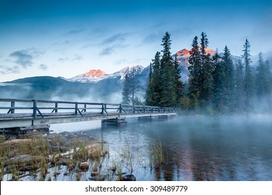 First glimpse of a golden sunrise on a misty and foggy morning at Pyramid Lake in Jasper National Park, Alberta, Canada. The wooden bridge leads to Pyramid Island on the lake.