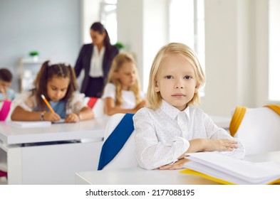 First Grader Elementary School. Portrait Of Cute Little Serious Boy Sitting At Desk During Lesson In School Classroom. Handsome Caucasian Blond Kid Boy Sitting Straight And Looking At Camera.