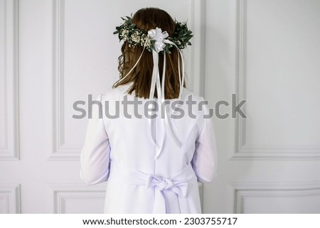 first Holy Communion. A girl in a white communion dress, standing with her back, Caucasian, shoulder-length dark hair. She has a beautiful wreath of fresh flowers on her head