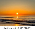 As the first light of dawn breaks over the horizon, a fiery orange sun rises from the azure waters of the ocean, painting the sky and waves with a breathtaking golden hue over Myrtle Beach, SC