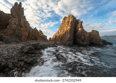 The first light of dawn rises over the rocky coastline of Quang Ngai, Vietnam, as waves gently crash against scattered stone outcrops. Golden sunlight reflects across the sea surface, creating a dramatic contrast between the rugged rocks and the soft morning sky. The rhythmic sound of the surf and the warm glow of sunrise capture the raw beauty of Vietnam’s central coast.