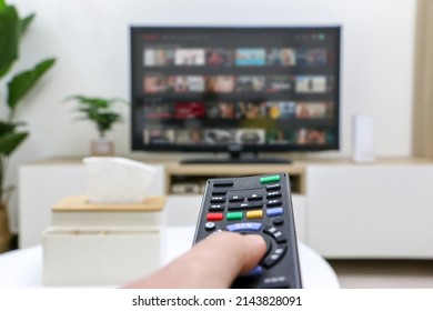 First Person Perspective Of Hand Holding Remote Control, Choosing What To Watch On Streaming Platform Displayed On Television Screen In Background But Out Of Focus, Depicting Home Life