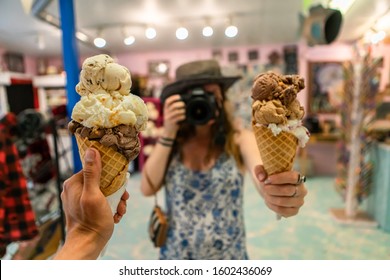 A First Person Perspective Of A Man Holding An Ice Cream Cone, In The Back Ground Out Of Focus A Woman Holds A Second Cone Holding A Camera