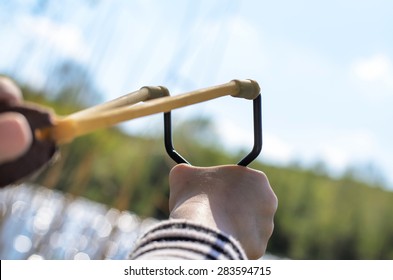 First Person Perspective Of Young Boy Holding Sling Shot, Pulling Rubber Strips Back In Anticipation Of Catapult Shot Across Lake