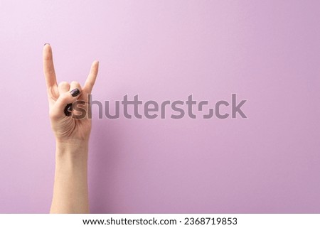 From first person top view, a black-manicured hand of a young woman creates the rock sign on lilac. Nonverbal language concept, space for text or advertisements