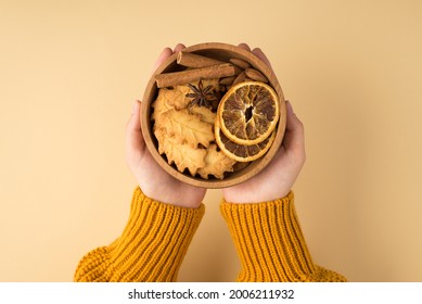 First person top view photo of woman's hands in yellow pullover holding wooden bowl with leaf-shaped cookies dried lemon slices and cinnamon sticks in palms on isolated light orange background