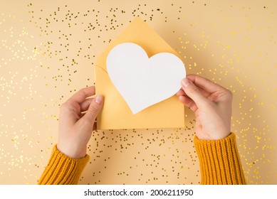 First person top view photo of female hands in yellow sweater holding open pastel yellow envelope with white paper heart over scattered golden sequins isolated light orange background with copyspace